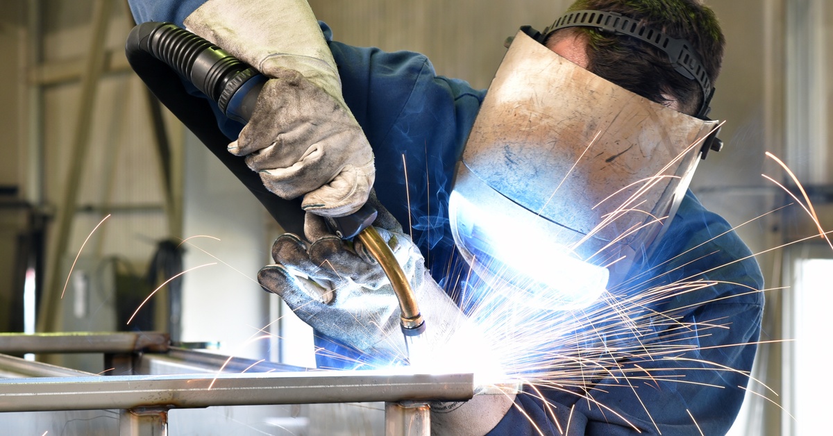 A man in blue protective overalls and a welding helmet, welding together the joints of thin pipes.