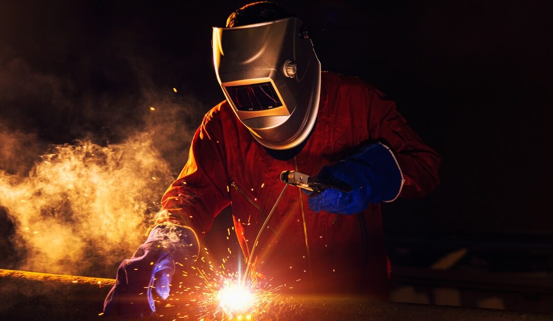 A worker in blue gloves, a welding helmet, and an orange protective suit, welding a pipe. The lighting is dynamic, black, and orange.