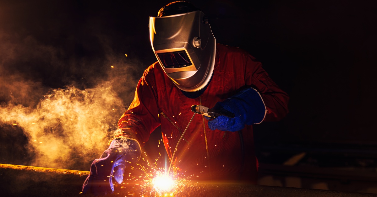 A worker in blue gloves, a welding helmet, and an orange protective suit, welding a pipe. The lighting is dynamic, black, and orange.