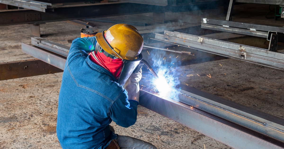 A worker in a yellow helmet and a denim jacket, kneeling down to weld a large metal structure.