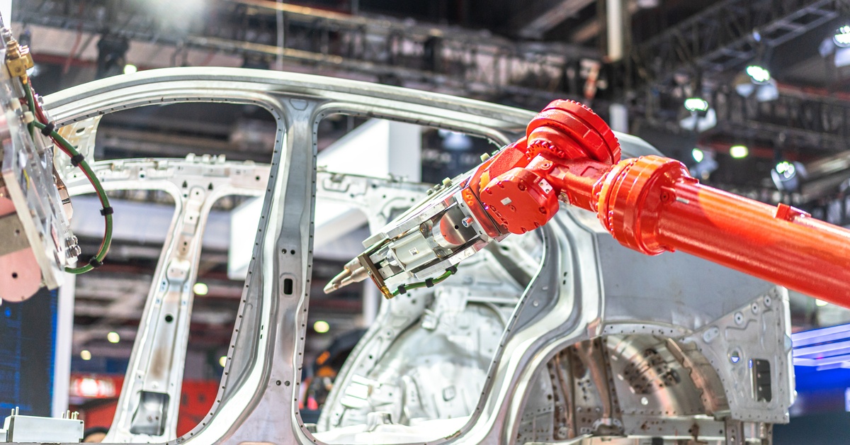 A singular red robot welding arm poised in front of the silver frame of a nondescript vehicle.