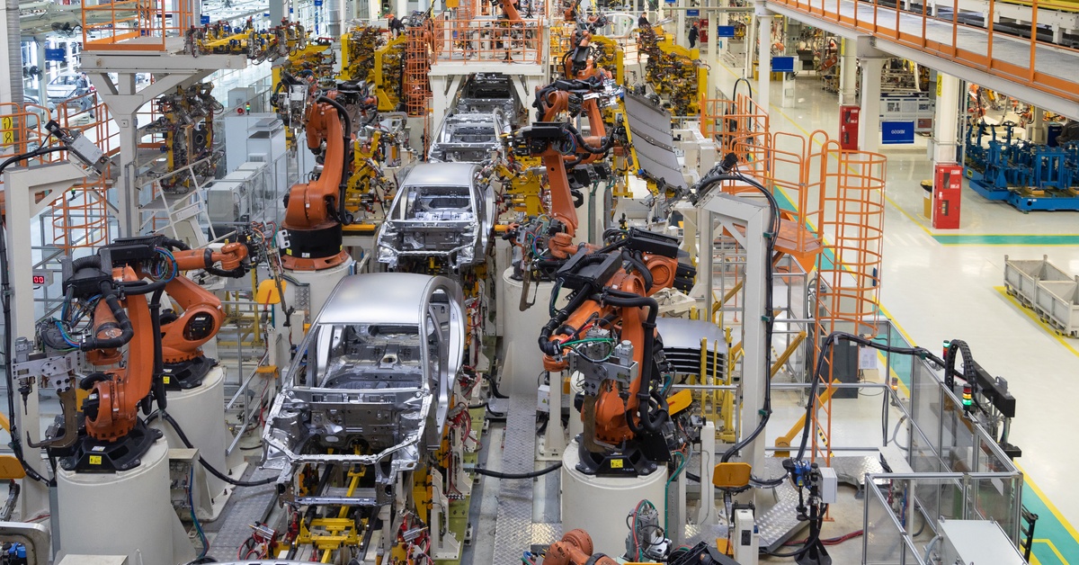 A high-angle shot of an automotive assembly shop with a line of car frames being worked on by robotic welding arms.