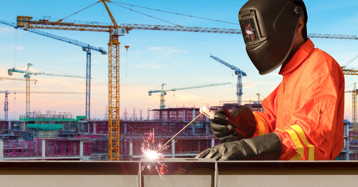 A person in an orange work suit, black gloves, and a welding helmet is welding a railing in front of a high-rise construction site.