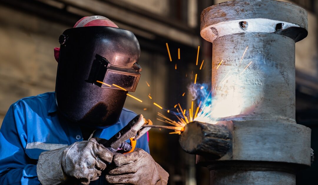 A person in a blue button-up shirt, a welding shield, and gloves, using stick welding on a large, vertical pipe.