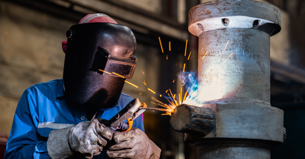 A person in a blue button-up shirt, a welding shield, and gloves, using stick welding on a large, vertical pipe.