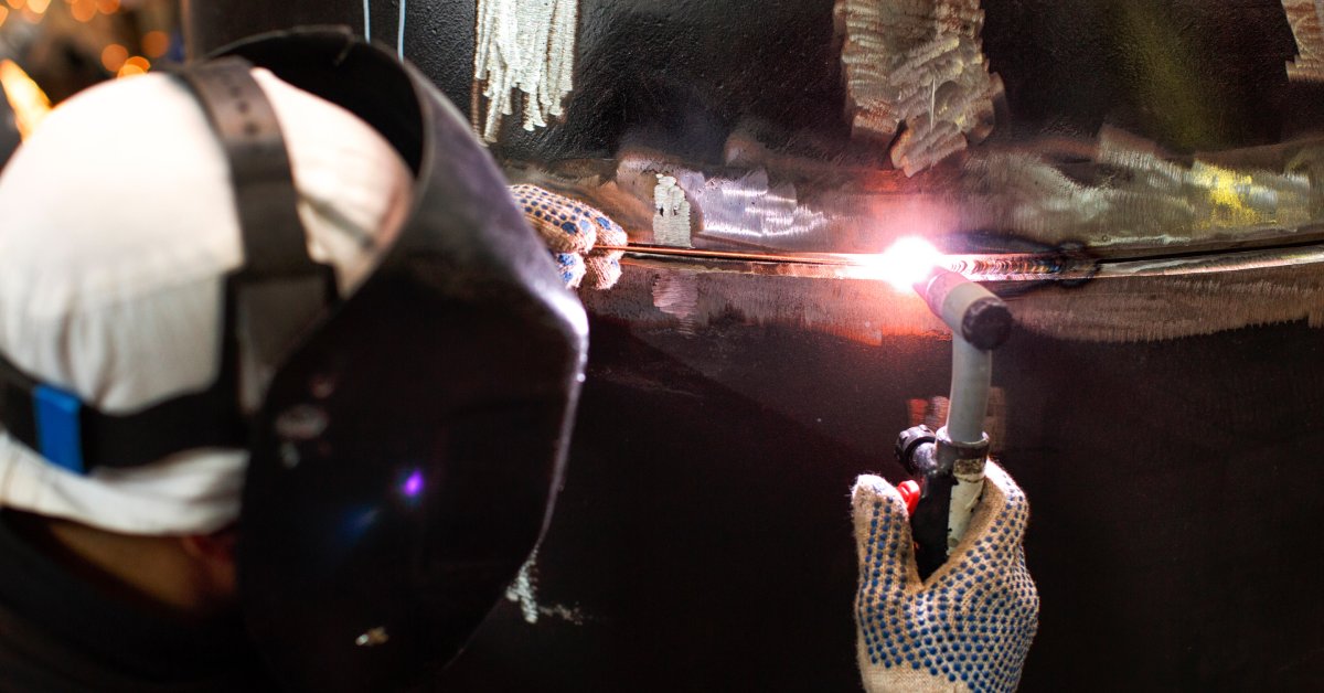 A welder in a white helmet and white gloves is welding a large steel pipe using gas tungsten arc welding.