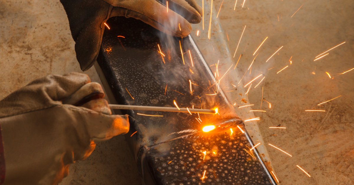 A close-up of a person wearing thick brown welding gloves, welding two long pieces of metal together.