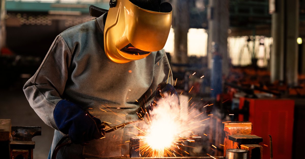 Sparks fly as a welder in a protective apron and yellow welding mask holds a torch to a material in a workshop.