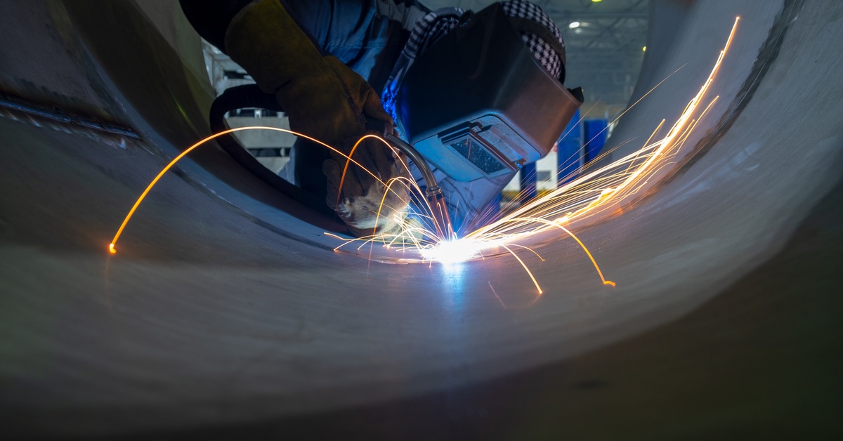 A welder working in a large pipe, wearing welding gloves and a protective helmet, using MIG. Sparks fly from the nozzle.