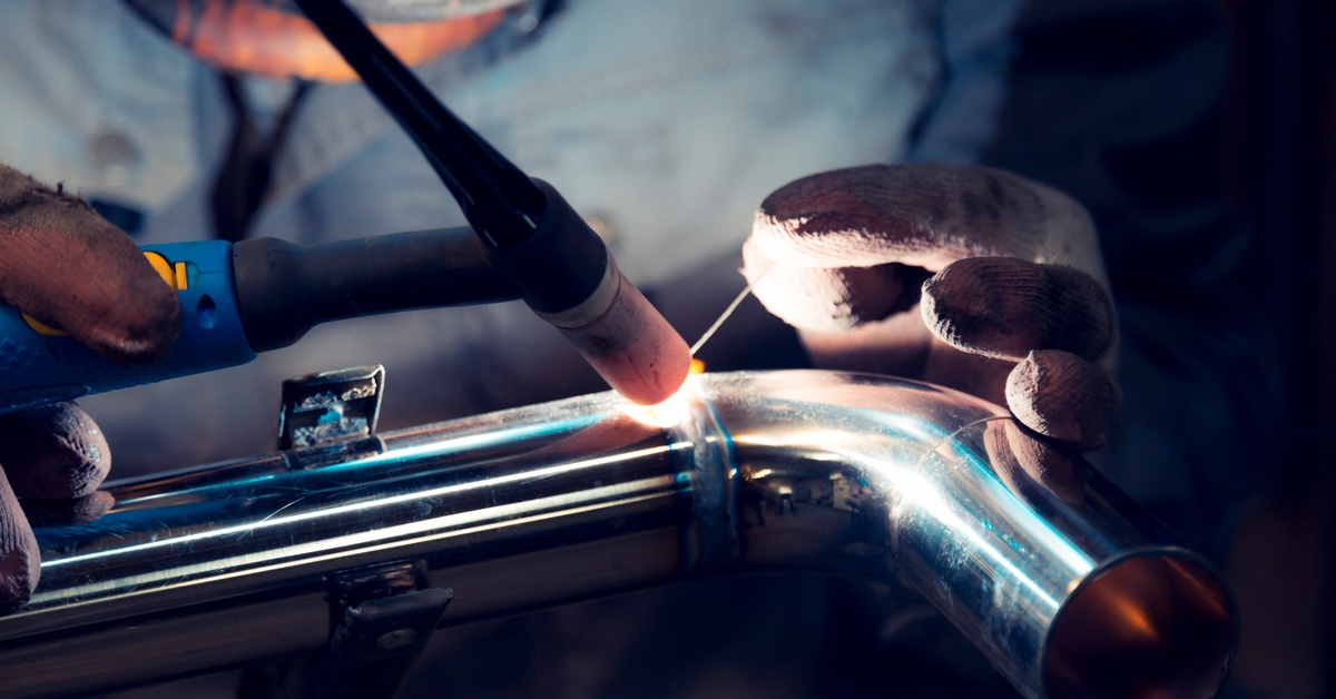 A close-up of a welder using TIG welding to create a fine seam on the curve of a silver pipe.