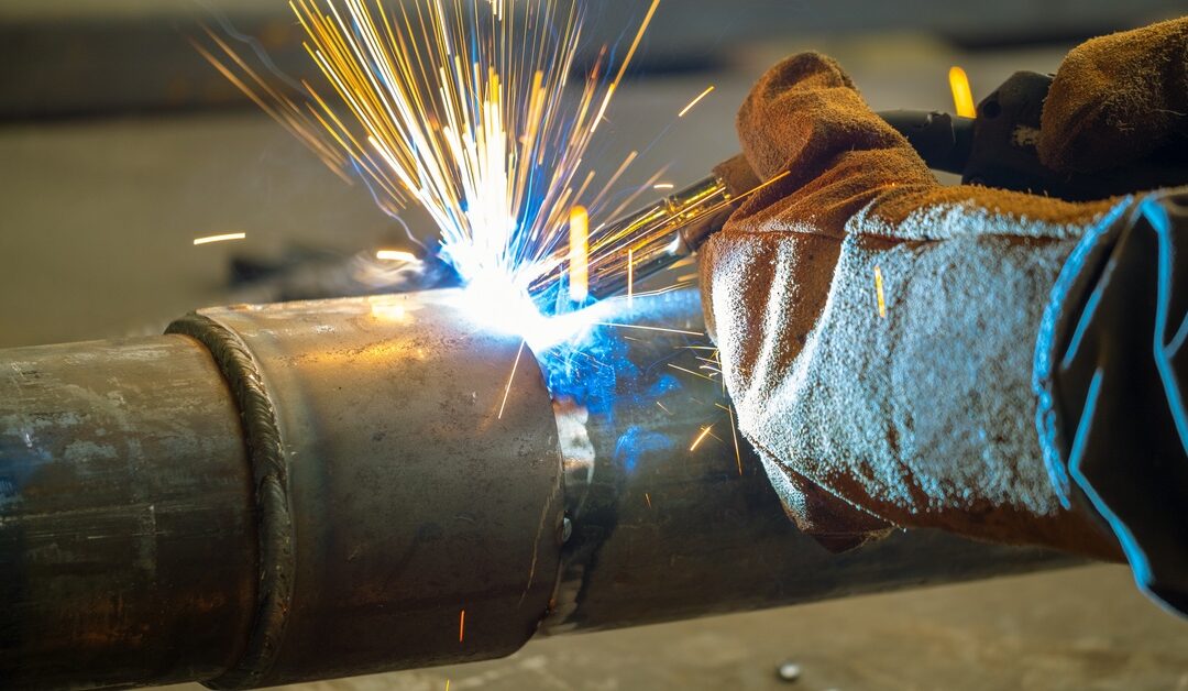 A close-up of a welder, wearing thick welding gloves, using arc welding to secure two pipe connections together.