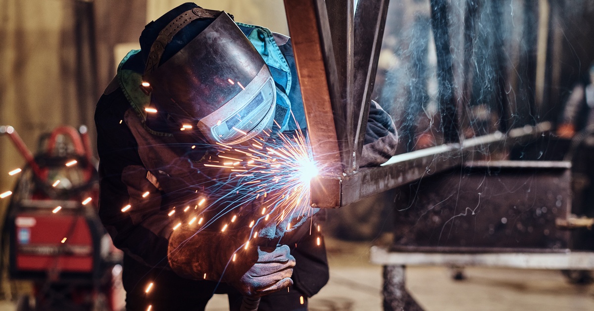 A welder in the proper safety gear—helmet, gloves, and overalls—welding a long piece of railing.