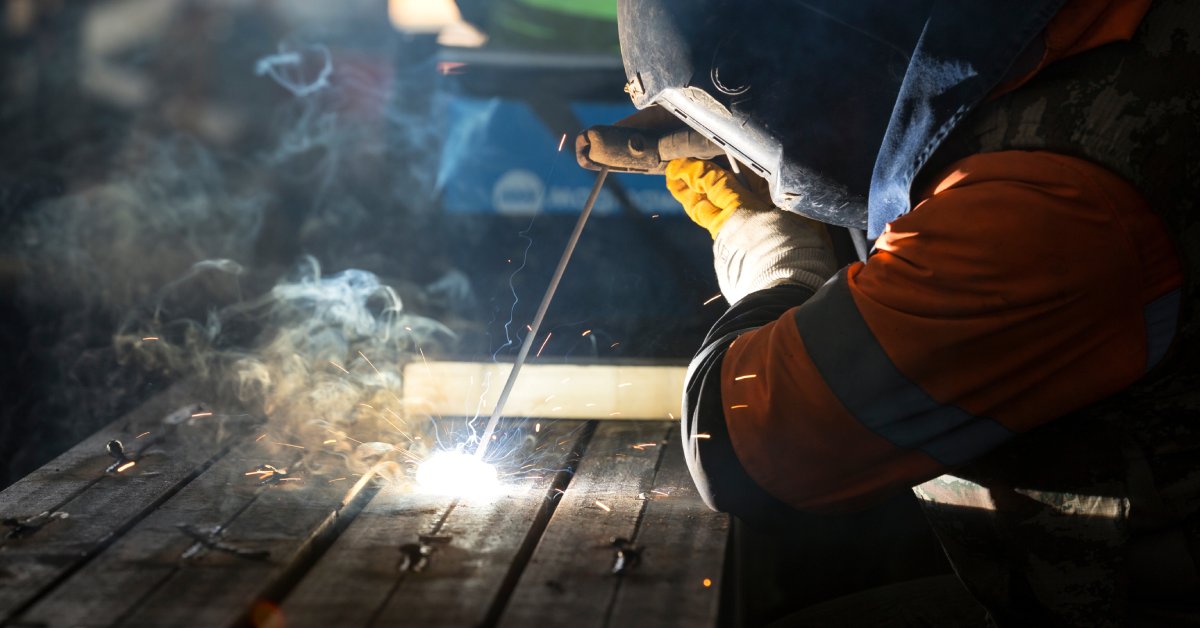A person in a welding helmet and orange protective suit, with the focus on the end of the sparking stick weld.