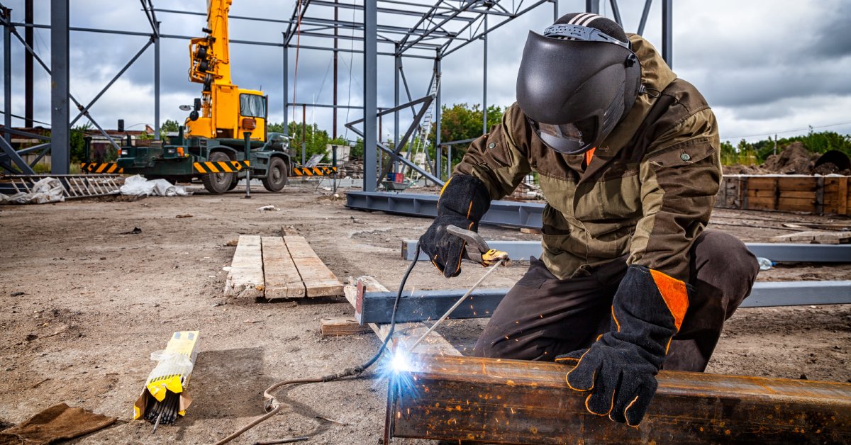 A welder wearing protective gear, kneeling down in a large construction site, using stick welding to weld the end of a beam.