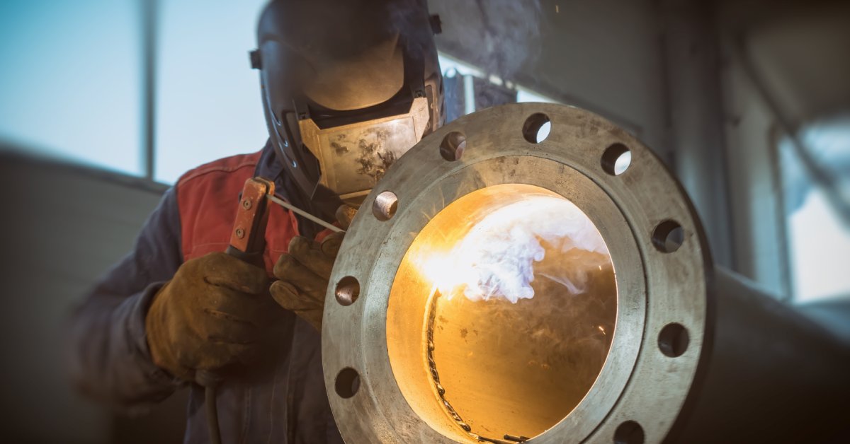 A person using stick welding to fuse a pipe flange to the end of a metal pipe, with the smoke emitting through the tube.