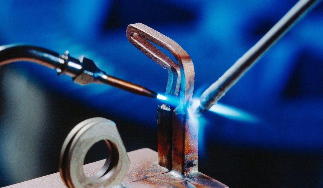 A copper component being brazed with a brazing rod and a soldering iron emitting a blue flame against a blue background.