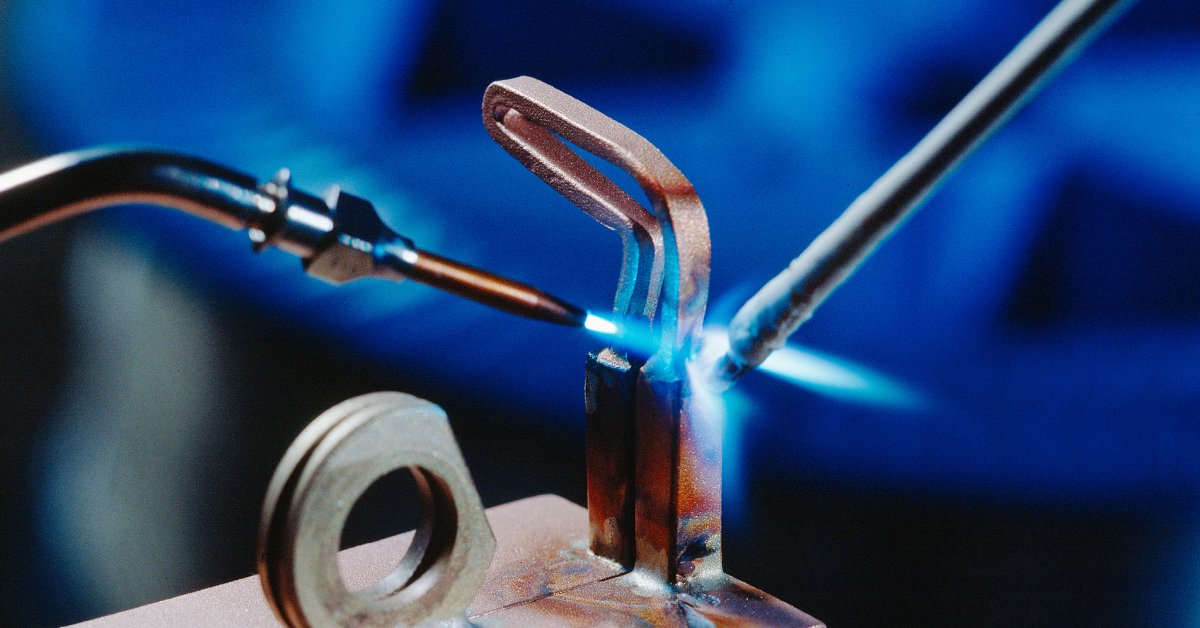 A copper component being brazed with a brazing rod and a soldering iron emitting a blue flame against a blue background.