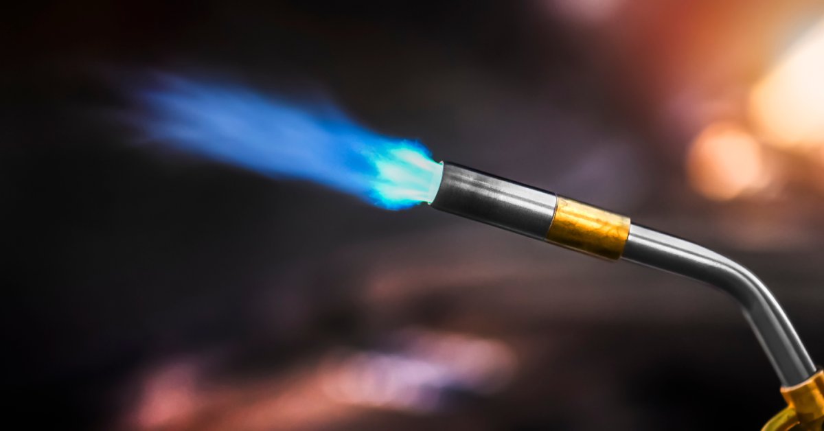 A gas torch soldering iron set against a dark, bokeh background, with a large blue flame spouting out.