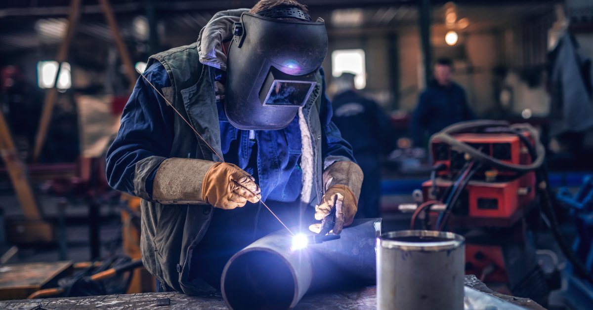 A person in full welding gear—suit, gloves, and mask—working in a workshop with a soldering rod over a large piece of metal.