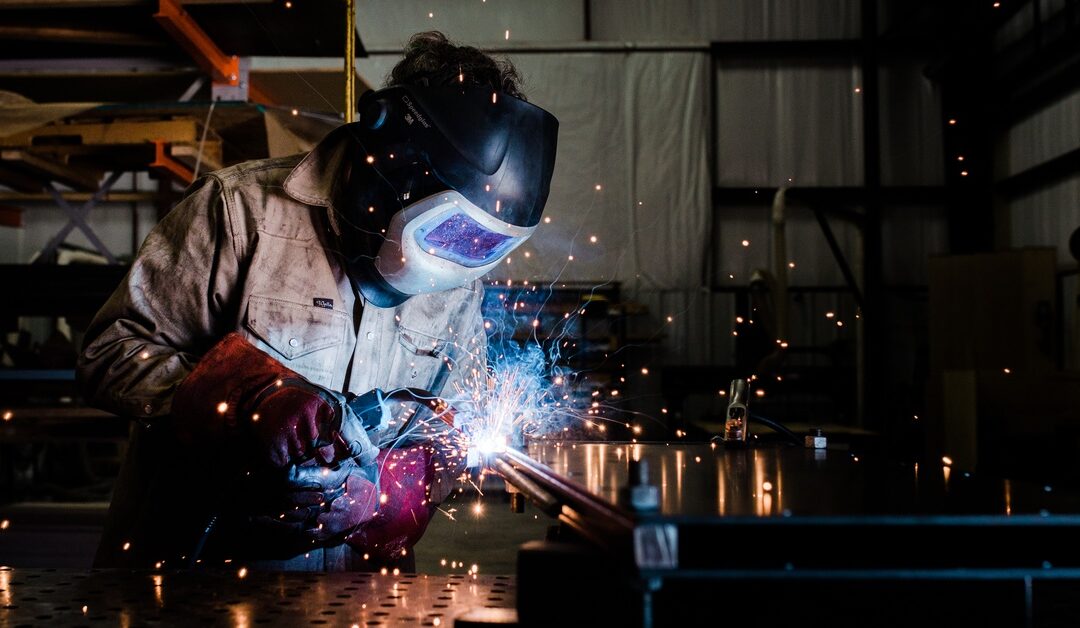 A person wearing a welding mask and jumpsuit using a torch to weld metal on a workbench in a workshop.