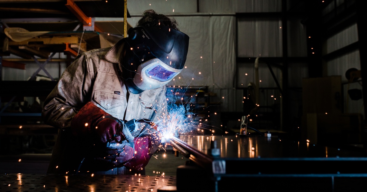 A person wearing a welding mask and jumpsuit using a torch to weld metal on a workbench in a workshop.