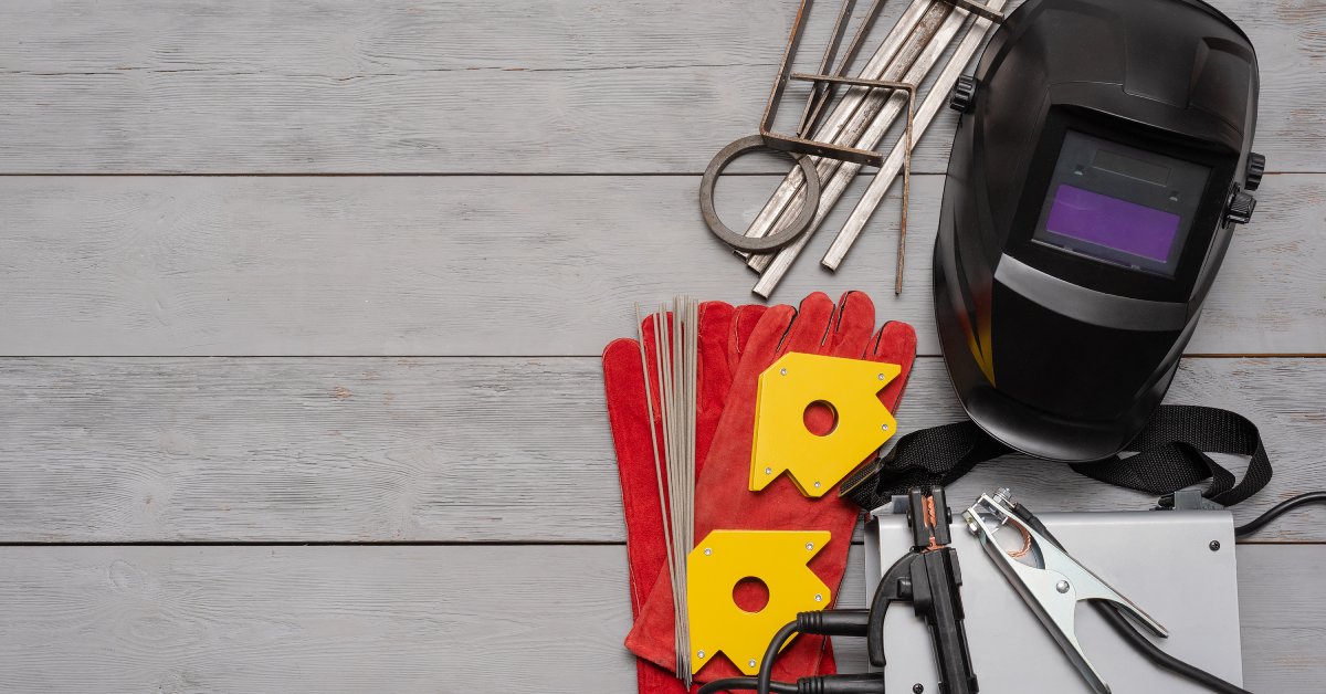 Various welding supplies, such as a helmet, welding rods, and gloves, placed on a gray wooden plank surface.