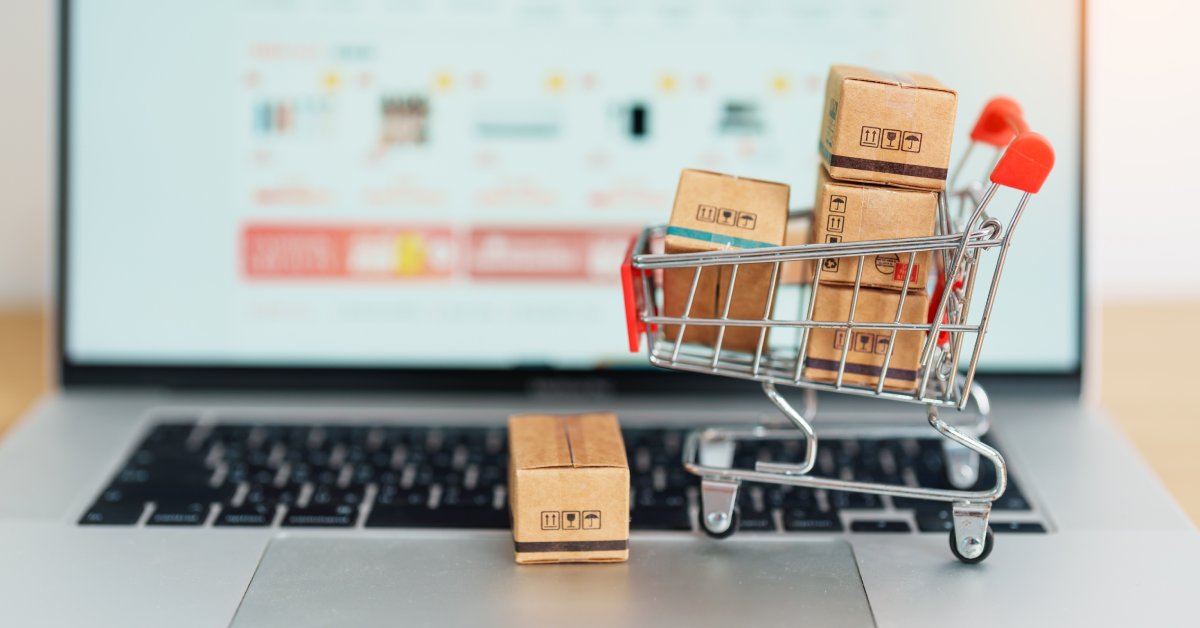 A laptop displaying a shopping website, while a model shopping cart filled with shipping boxes rests on the laptop itself.