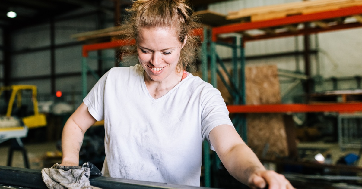 A woman with her hair in a bun wearing a white t-shirt smiling as she cleans a piece of metal with a cloth in a warehouse.