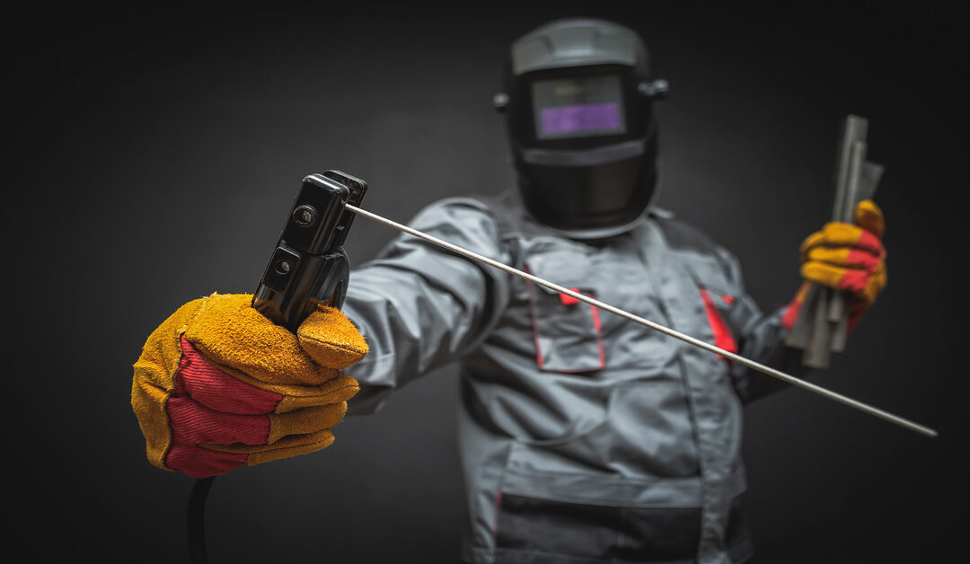 A welder wearing full protective gear, including a jumpsuit and helmet, and holding welding terminals.