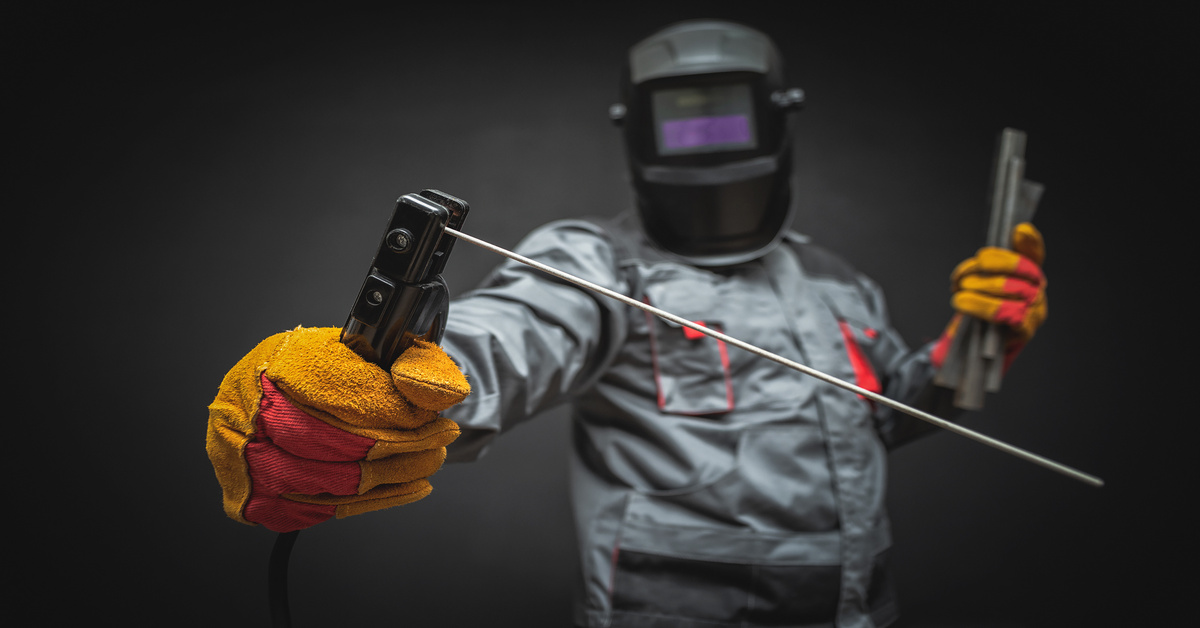 A welder wearing full protective gear, including a jumpsuit and helmet, and holding welding terminals.
