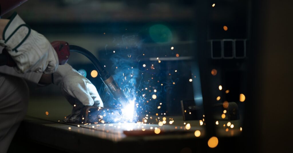 A worker performing welding in a factory while wearing protective gloves, with bright sparks flying from metal.