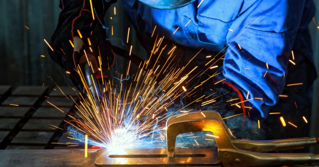 A close up of a man wearing a welding mask while using a torch on metal with light from the weld visible.