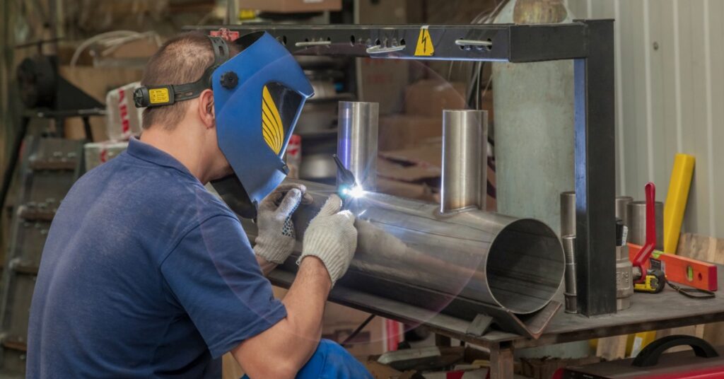 A man kneeling beside a large pipe and welding along the seam while the pipe is held in place in a bright room.