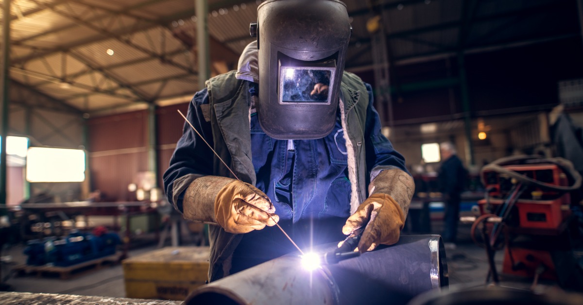 A masked welder working on metal at a workshop table while other workers stand blurred in the background.
