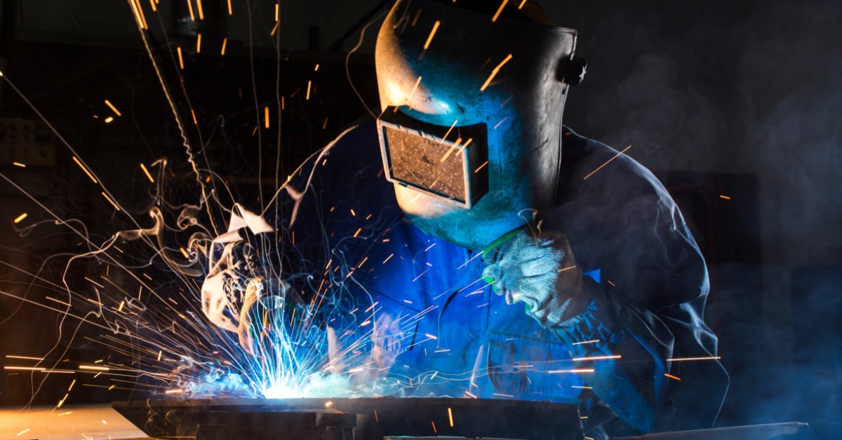 A person wearing a protective welding helmet using a torch on metal, with bright arc light and sparks.