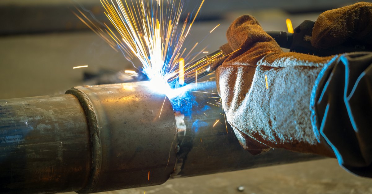 A gloved person welding a clamped pipe connection, with blue arc light and sparks near the metal joint.