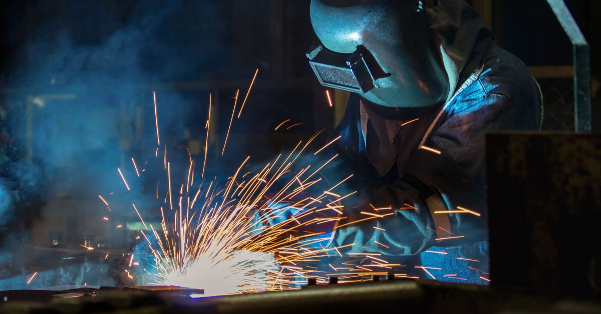 A person wearing a welding helmet working beside bright sparks and blue smoke inside a dark industrial workspace.
